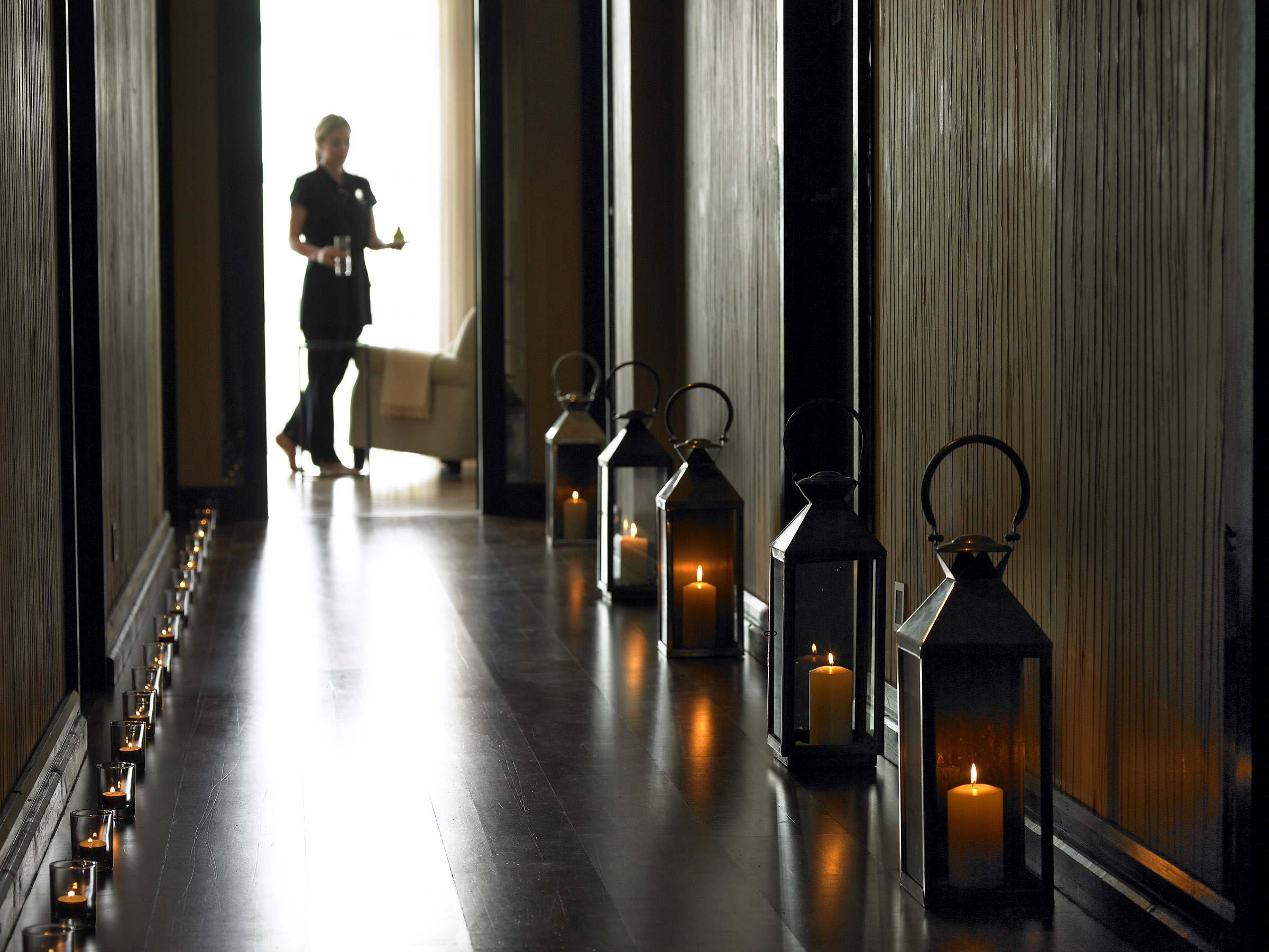 Dimly lit hotel hallway with candle-lit lanterns and a staff member carrying refreshments, leading to a cosy lounge area.