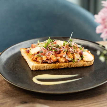 Elegant open sandwich with herbs and drizzle, paired with white wine on a chic dining table setting.