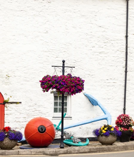 Charming white cottage with vibrant red door, colourful hanging baskets, and nautical decor in a quaint coastal village.