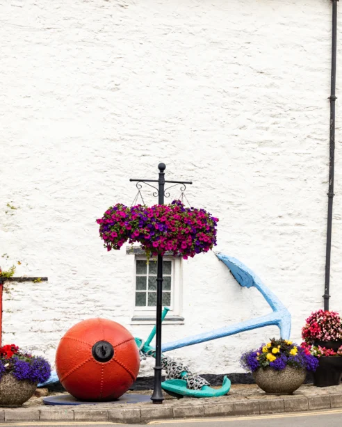 Charming coastal hotel exterior with colourful flowers, rustic red door, nautical decor, and inviting village atmosphere.