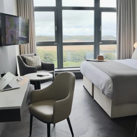 Elegant hotel room with king bed, scenic countryside view, cosy seating area, flat-screen TV, and sleek workspace.