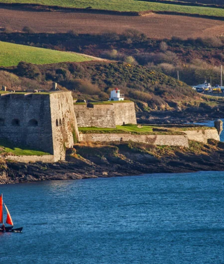 Stunning sea view of historic coastal fort with a vibrant red sailboat, lush greenery, and nearby marina.