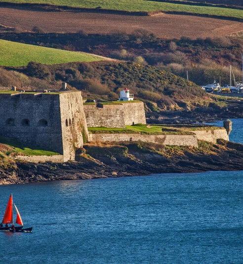 Stunning sea view of historic coastal fort with a vibrant red sailboat, lush greenery, and nearby marina.